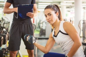 Young athletic girl lifting weights while her personal trainer monitoring her workout in gym.