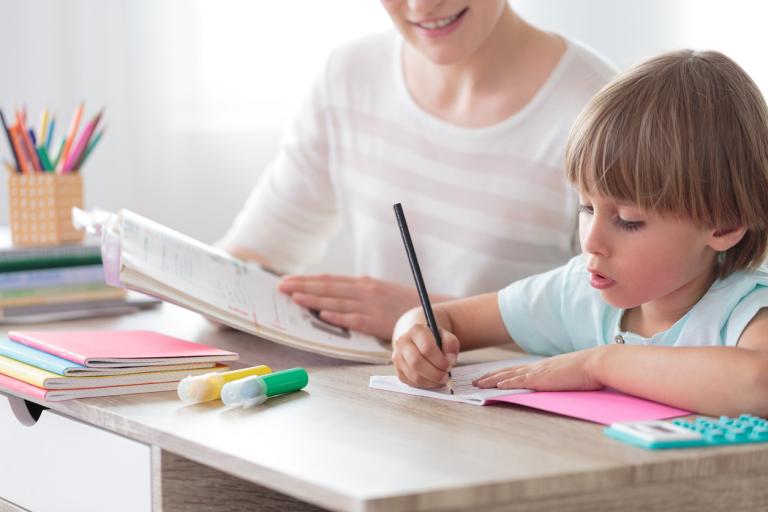 Boy focusing on homework while sitting with mother at desk with notebook and colored pens.