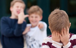 An upset elementary school boy hides his face while being bullied by two other boys as an example of a negative learning environment. Shot in front of their elementary school.