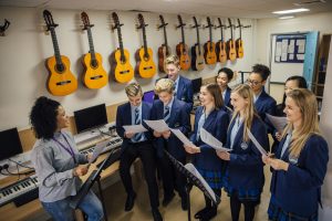 Class of teens are in their music lesson at private schools. There is a female teacher and the class are practicing choir with her.