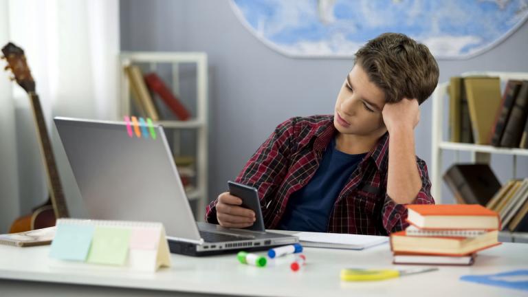 Young student browsing his cellphone while doing homework to suggest time blindness.