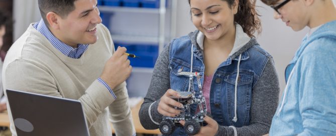 Diverse male and female high school students build a robot in technology class. A male teacher is helping them. Robot parts and a laptop are on the table.