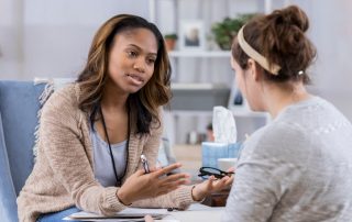 Serious female counselor gestures while talking with young female student about Hurricane Harvey. The counselor is holding eyeglasses and a pen.