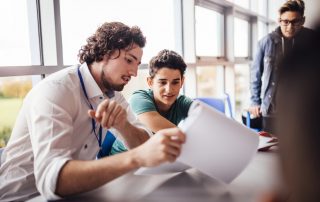 Teacher giving student customized instruction. The student looks happy as they sit side by side at a table. Another student looks on.