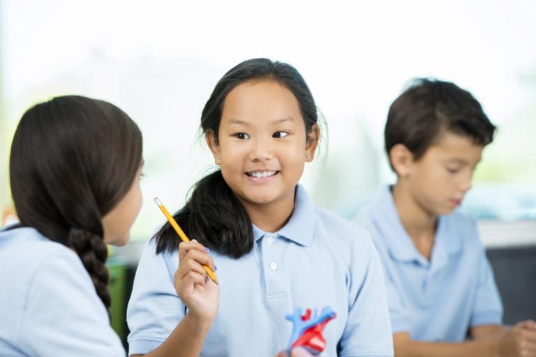 A confident, school age Asian female student talks to her Hispanic classmate. International students work together on a science project in class. They are studying a model of the human heart at a private school