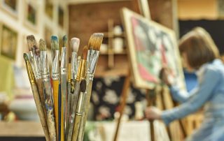 Set of paintbrushes in the foreground and a girl painting a picture on the easel in art studio on the background