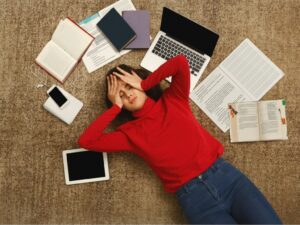 Tired student dealing with homework stress lying on the floor with books and gadgets.