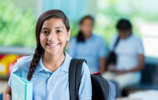 Confident international student before class. She is standing in her classroom. He has a braid and is wearing a backpack and a school uniform. Students are in the background talking.