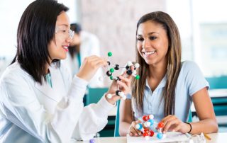 Young Asian female teacher helps teenage private high school student study molecular structure from a customized curriculum. The teacher is holding a plastic educational model. The teenage girl is interested as her teacher talks.