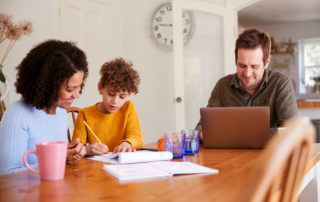 Father Works On Laptop As Mother Helps Gifted & Talented Child With Home work On Kitchen Table