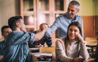 Happy mid adult teacher giving a friendly handshake to teenage boy on a class. Focus is on teacher. Tailored learning concept