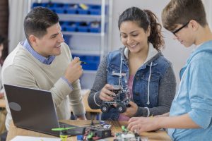 Diverse male and female high school students build a robot in technology class. A male teacher is helping them. Robot parts and a laptop are on the table.