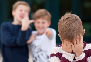 An upset elementary school boy hides his face while being bullied by two other boys as an example of a negative learning environment. Shot in front of their elementary school.