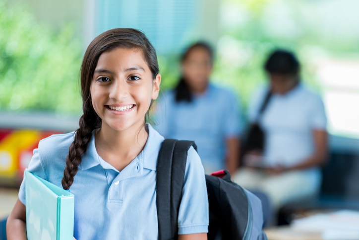 Confident international student before class. She is standing in her classroom. He has a braid and is wearing a backpack and a school uniform. Students are in the background talking.