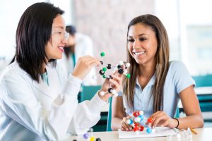 Young Asian female teacher helps teenage private high school student study molecular structure from a customized curriculum. The teacher is holding a plastic educational model. The teenage girl is interested as her teacher talks.