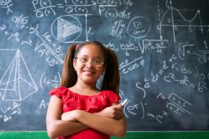 Concepts on blackboard at school. Young people, students and pupils in classroom. Smart hispanic girl writing math formula on board during lesson. Portrait of female child smiling, looking at camera
