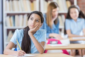 Cute Hispanic middle school girl looks away while daydreaming in class. Struggle with writing concept