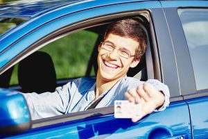 Teenager wearing glasses and jean shirt sitting behind wheel and holding out his driver's license through car window