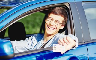 Teenager wearing glasses and jean shirt sitting behind wheel and holding out his driver's license through car window