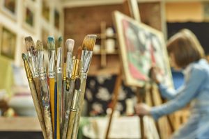 Set of paintbrushes in the foreground and a girl painting a picture on the easel in art studio on the background
