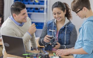 Diverse male and female high school students build a robot in technology class. A male teacher is helping them. Robot parts and a laptop are on the table.