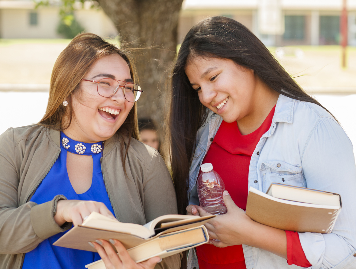 Multi-ethnic group of high school girls talking together outdoors. The Hispanic and Native American group of girls are holding textbooks and backpacks. The school building is in the background. Another girl is sitting by tree. International students