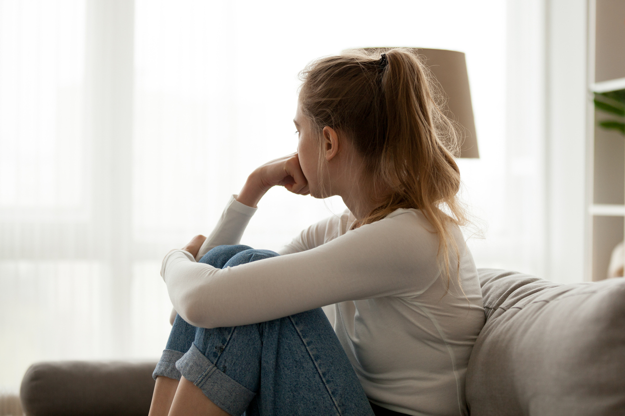 Side view young teen with anxiety and depression looking away at window sitting on couch at home.