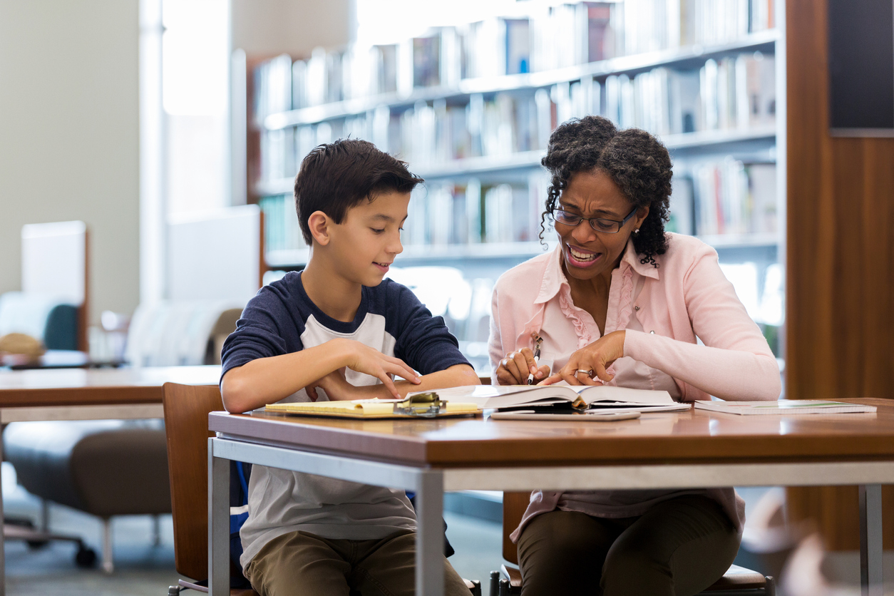 A middle school age boy sits with his mature female teacher using Personalized Learning Programs in the school library. They both look down as she reads from the textbook.