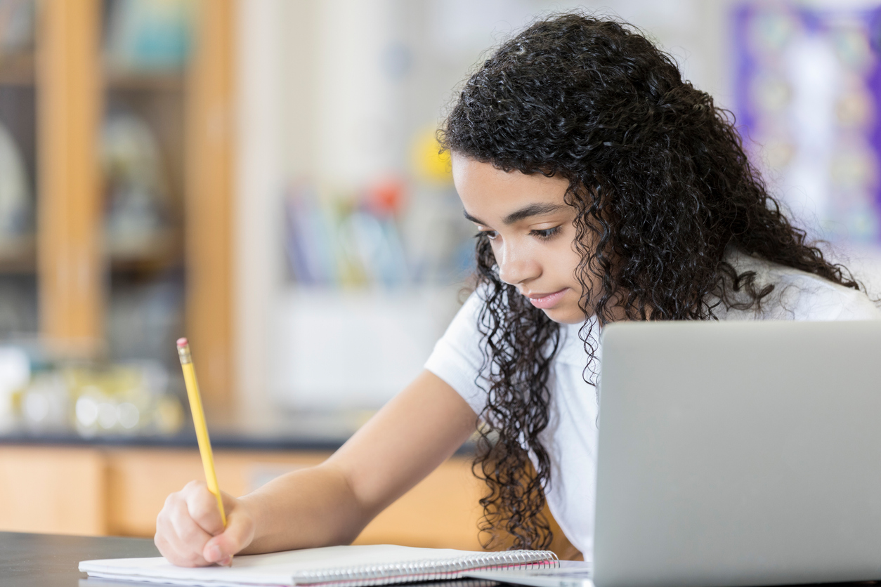 A serious middle school aged girl with Dysgraphia sits at a table in her science lab and looks down as she writes on a note pad. There is also a laptop computer on the table.