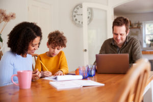 Father Works On Laptop As Mother Helps Gifted & Talented Child With Home work On Kitchen Table